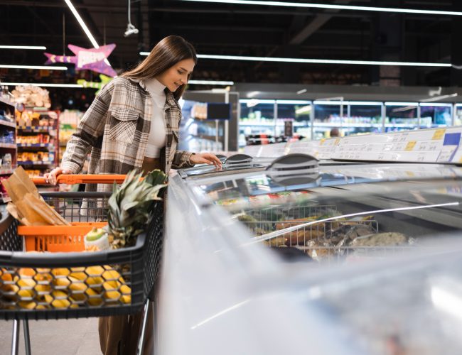 Young pretty woman with shopping trolley choosing products in frozen food department at supermarket. Shopping girl